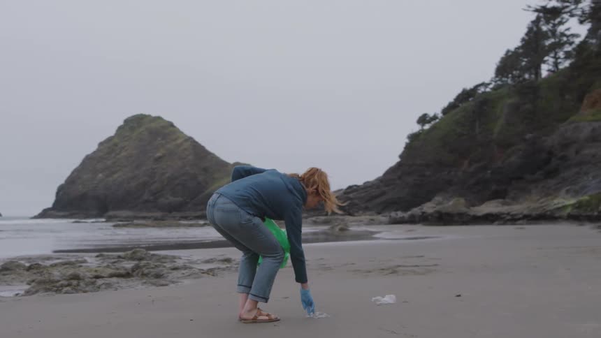 Woman picks up litter on beach with gloves and green bag surrounded by cliffs and trees under cloudy sky