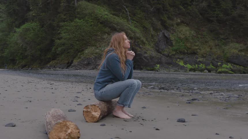 A young woman sits barefoot on a log of wood by the riverbank at midday the sunlight glinting off the water as she relaxes in the natural setting