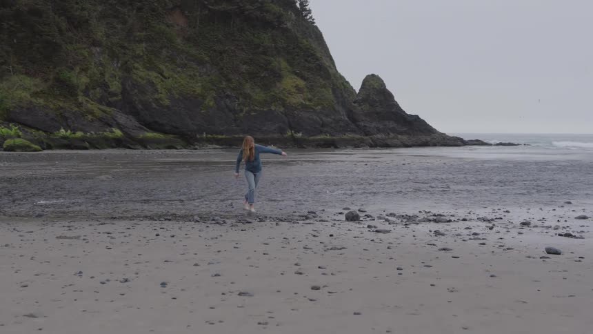 A woman wearing jeans walks across shallow water on a rocky shoreline at midday sunlight glinting off the stones and waves around her