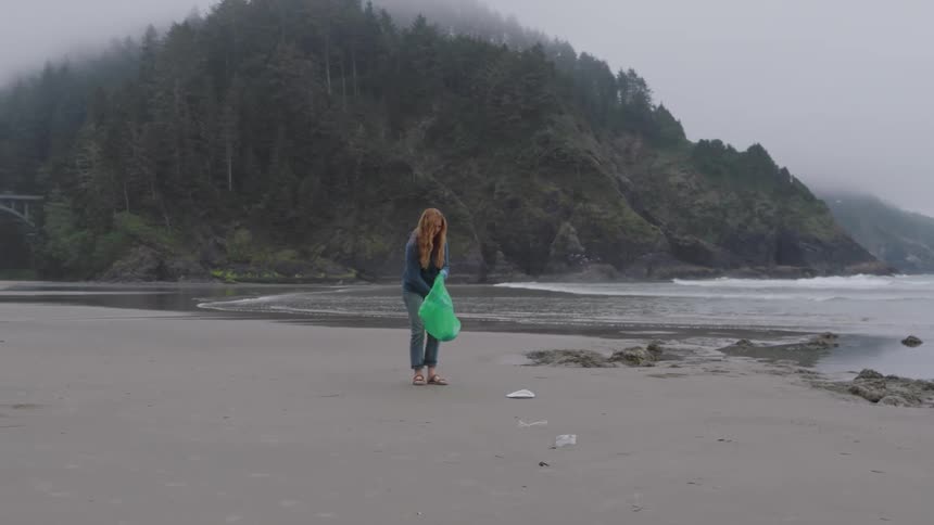 A woman with long red hair wearing jeans and a denim jacket cleans a coastal beach by picking up trash into a green bag set against a backdrop of a massive fogcovered treelined cliff and a large stone arch bridge