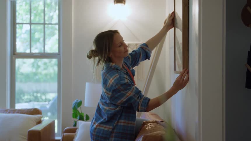 Young adult woman wearing a plaid shirt adjusting a frame hanged on the wall indoors during day time