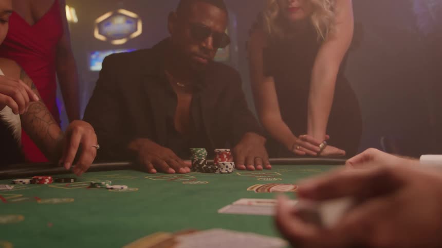 A black adult male wearing sunglasses and a suit is looking forward while gathering chips at a greenfelt gambling table surrounded by other people in a dimly lit casino