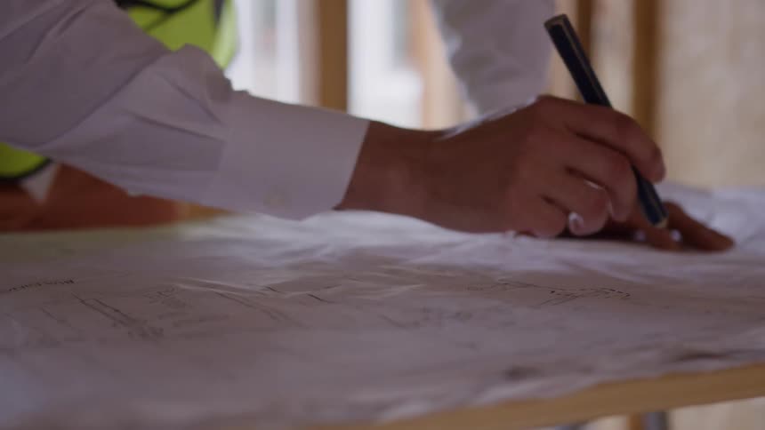 Person reviews architectural blueprint at construction site with wooden framing in background