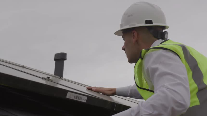 A worker wearing a hard hat and safety vest inspects a structure on a roof
