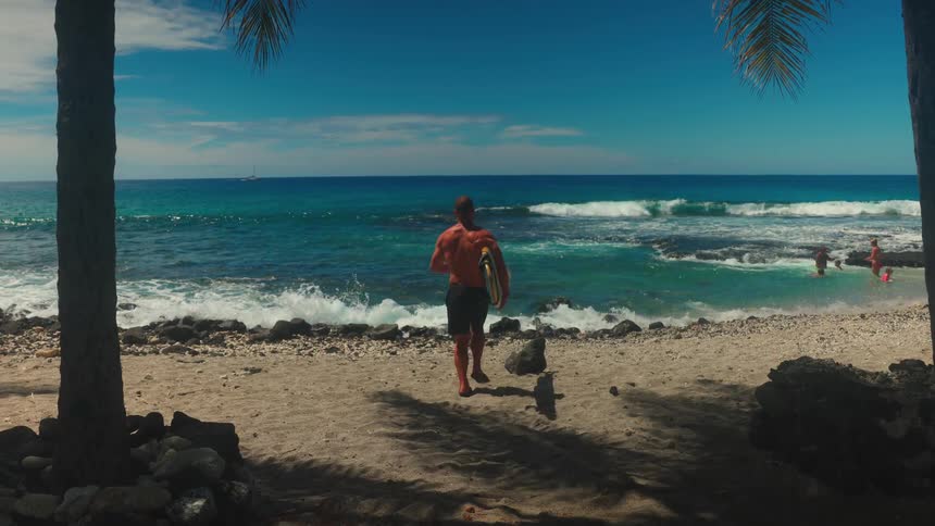 A shirtless man in dark shorts carrying a board walks toward the turquoise ocean water across a sunny sandy beach framed by palm trees as waves crash and people swim on the beach