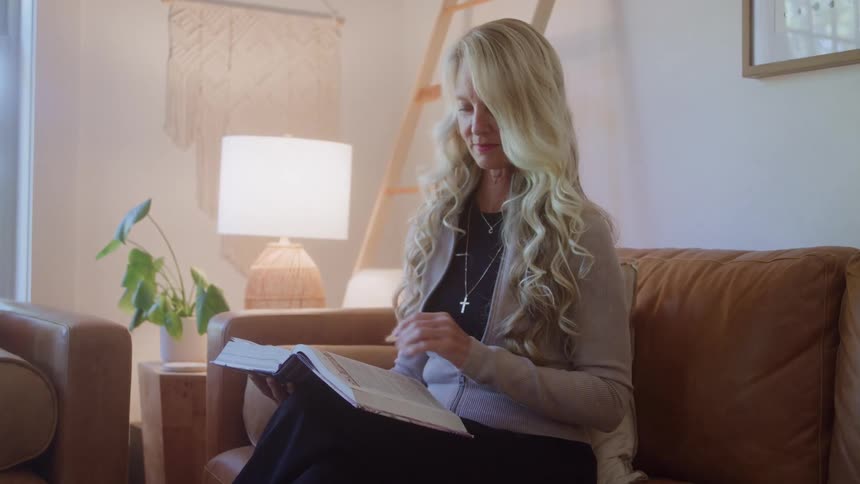 Woman reads book seated on leather couch in room lit by natural light