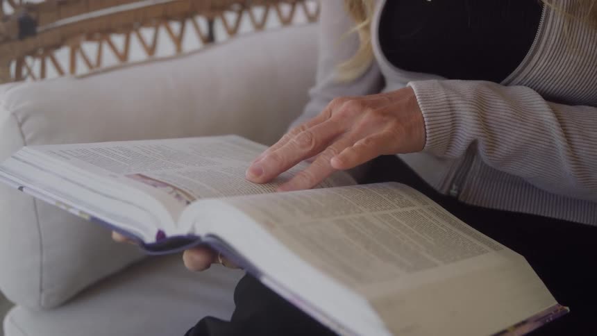 A closeup view shows a person sitting on a white couch reading a large book with textfilled pages using a finger to track the lines while concentrating on the material