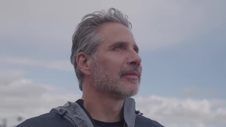 A closeup profile shot of a middleaged man with grey hair and a short beard wearing a dark jacket looking off into the distance or up towards a light blue cloudy sky
