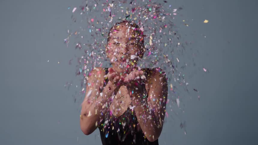 Portrait of a young black woman blowing confetti from her palms while sitting indoors in daytime