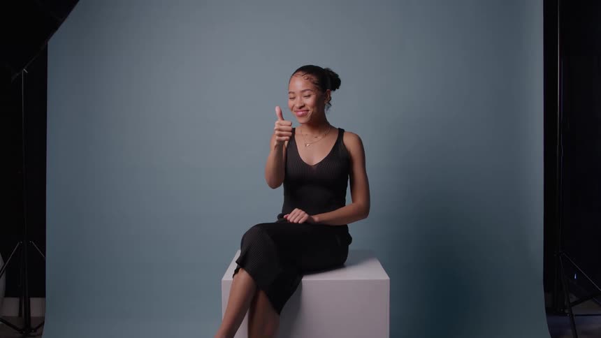 A young woman in a black knit dress sits on a white cube against a solid bluegrey backdrop in a studio smiling confidently while giving a thumbsup gesture to the camera
