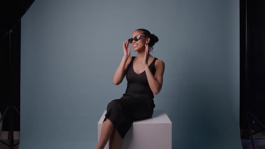 Young black woman wearing a black dress sitting and posing while adjusting sunglasses over her face at a studio indoors