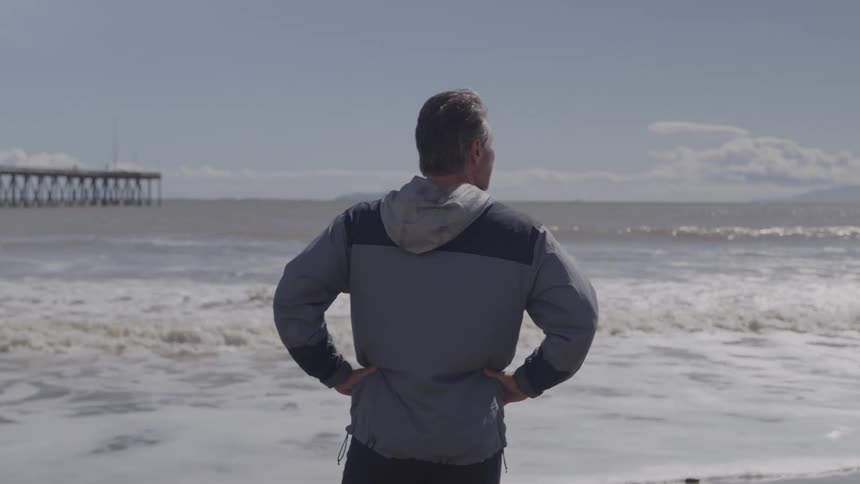Man stands on the beach with his hands on his hips watching waves roll in to shore
