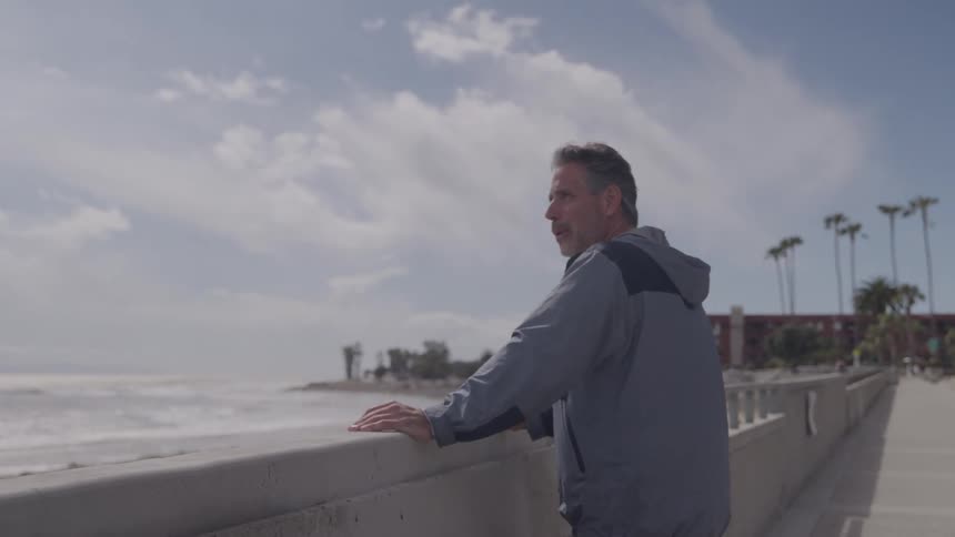 A middleaged man in a grey jacket leans against a concrete railing on an oceanfront walkway looking intently at the choppy water and cloudy horizon with palm trees and buildings visible in the distance