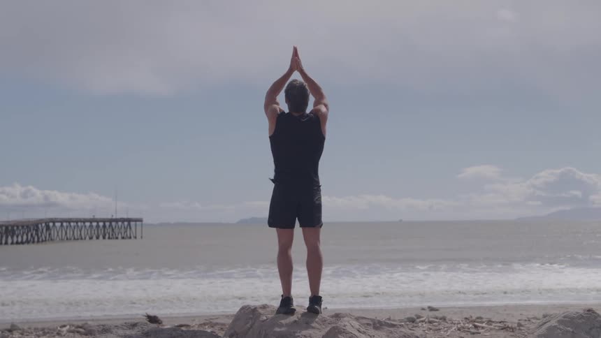 A man in black workout attire stands with his back to the camera on a rocky beach raising and lowering his arms in a stretching motion toward the ocean waves and a distant wooden pier under a cloudy sky