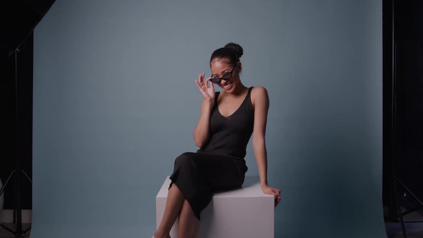 A young woman in a black knit dress sits on a white cube against a bluegrey studio backdrop smiling and laughing while playfully adjusting her dark sunglasses