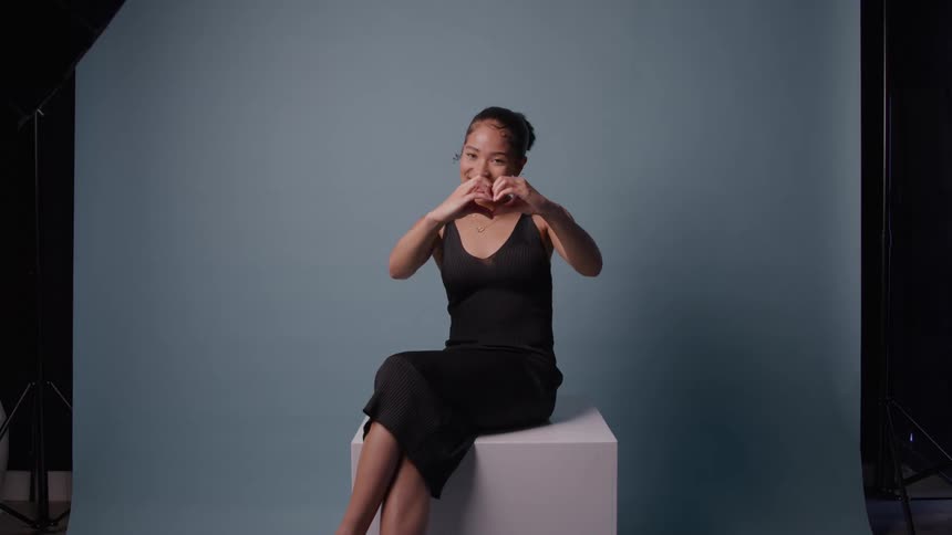 Woman sitting on a white cube forming a heart shape with her hands