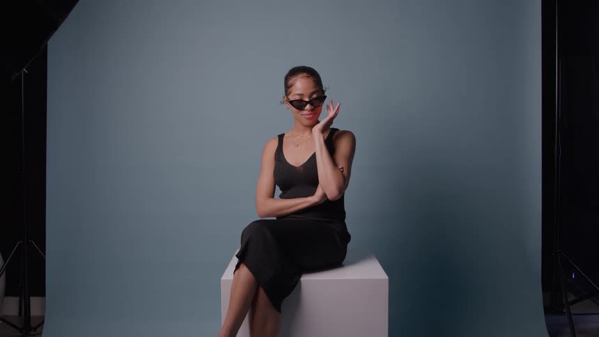 Woman sits on white cube adjusting sunglasses against blue backdrop in photo studio