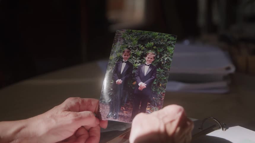 Hands hold photo of two smiling boys in black tuxedos standing before green bush