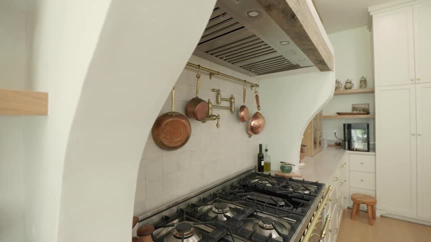 Stylish kitchen blends multiburner stove with copper cookware and brass accents beneath rustic hood