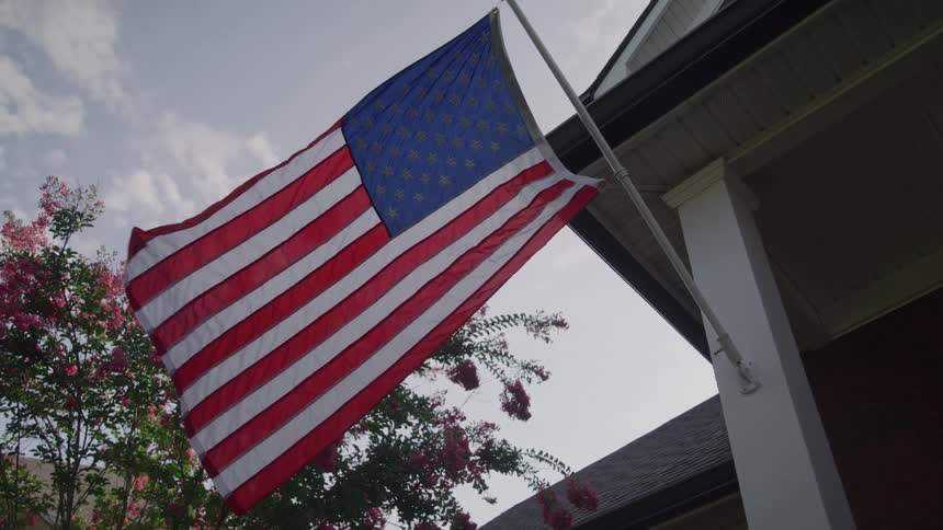 American flag waves proudly from house front framed by pink blossoms and partly cloudy sky