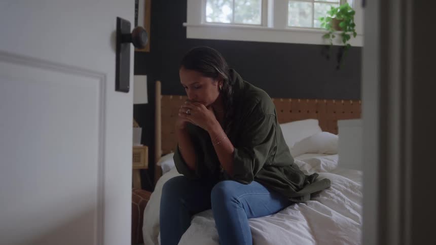 Woman sits at the edge of the bed in a contemplative moment softly lit by window light