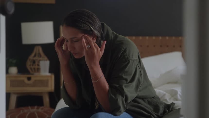 Woman sits at the edge of the bed with hands on temples in a quietly reflective moment