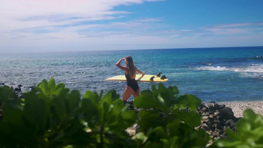 Woman with long hair holds yellow surfboard facing the sea on a rocky beach