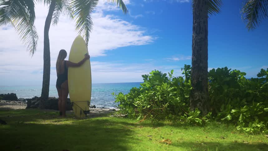 Woman stands on the beach holding her surfboard watching waves roll in to shore