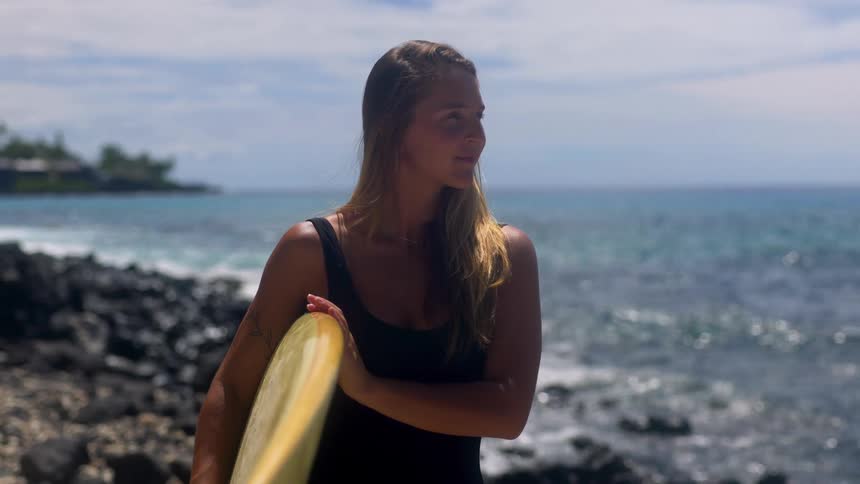 Woman with loose hair holds yellow surfboard on rocks facing the rough sea
