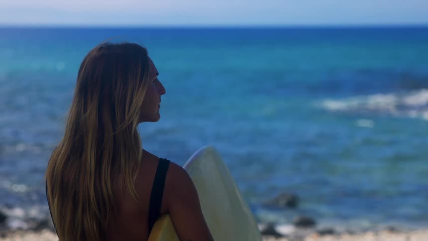 Woman stands on the beach holding her surfboard watching waves roll in to shore