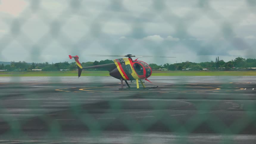 View through a fence of a helicopter sitting on the tarmac with the propeller spinning