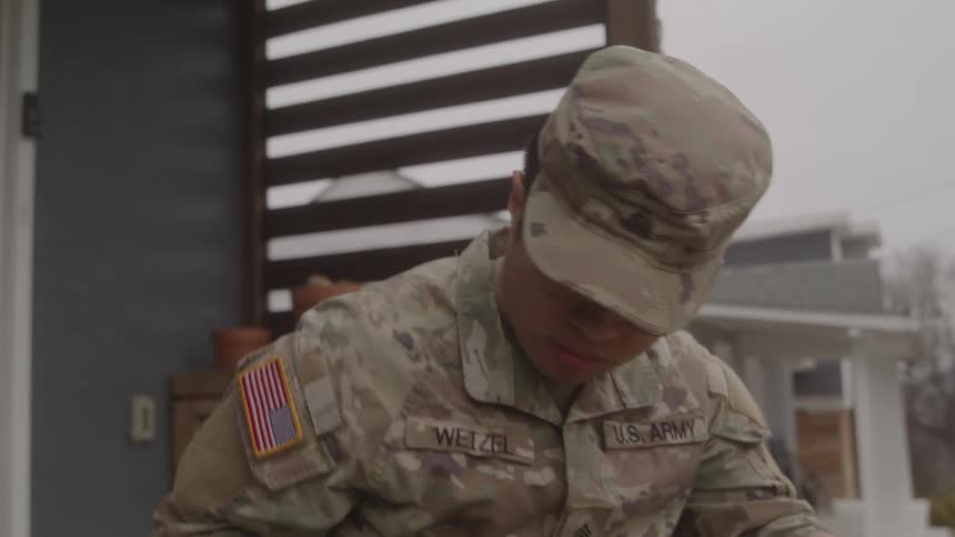 A military officer sits quietly on a patio at midday sunlight casting sharp shadows across the yard he places his hat firmly on his head picks up his backpack with practiced ease and strides away from the front yard with purpose