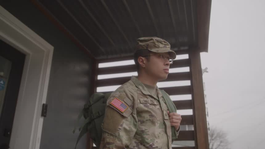 Service member with backpack and camouflage uniform stands by wooden wall under metal awning