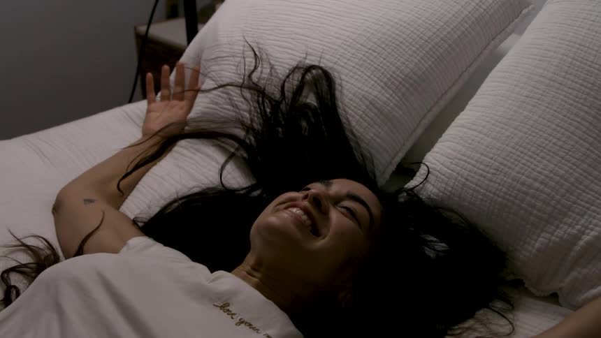 Woman with dark hair smiles lying on bed with arms spread over white quilt