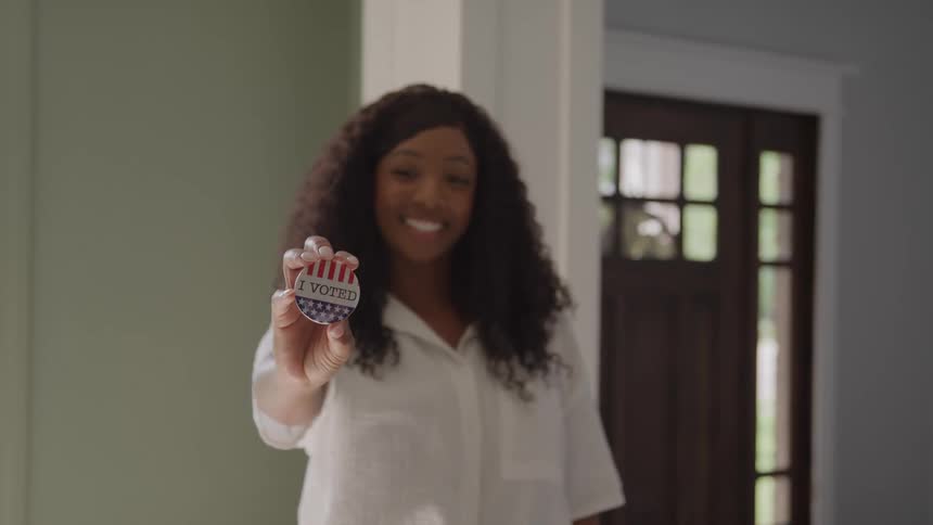 Woman smiles standing while holding circular sticker with i voted text in front of glass door