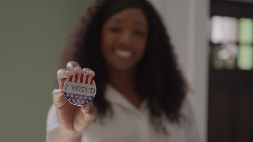 Person holds circular badge with patriotic design and i voted text indoors