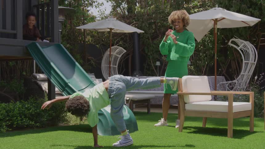 A child performs acrobatics on the backyard lawn under the sunny sky while the brother watches and cheers with excitement nearby the little sister sits on a slide observing the playful scene