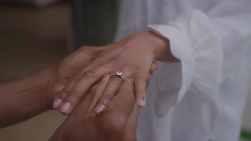 Hand places diamond ring on finger of person wearing white longsleeved ruffled blouse