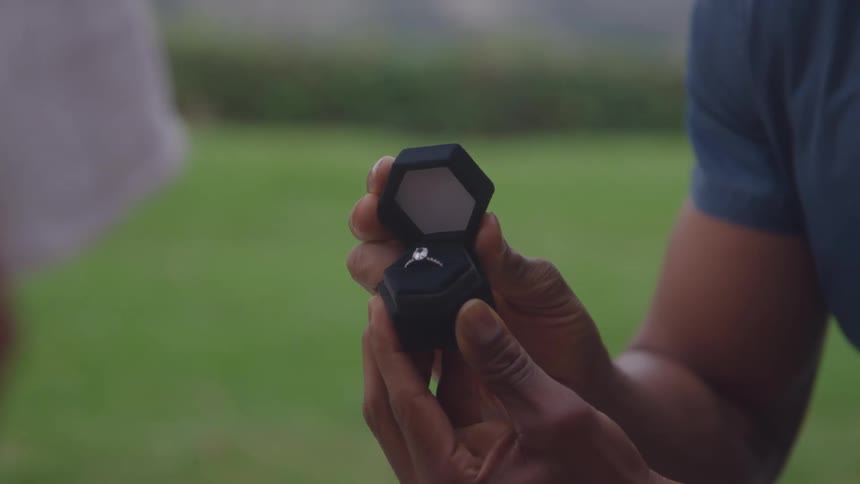 Person holds open black hexagonal box with diamond ring in grassy outdoor setting