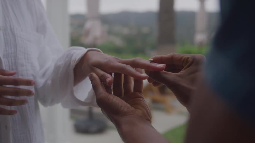 Hand placing a ring on the finger of a person dressed in white with blurred greenery and buildings in the background