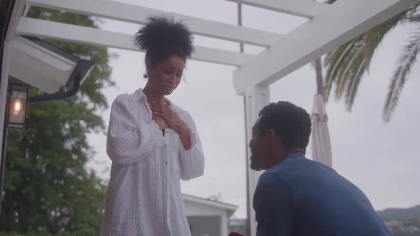 Man kneels holding ring box before emotional woman under white pergola in tropical setting