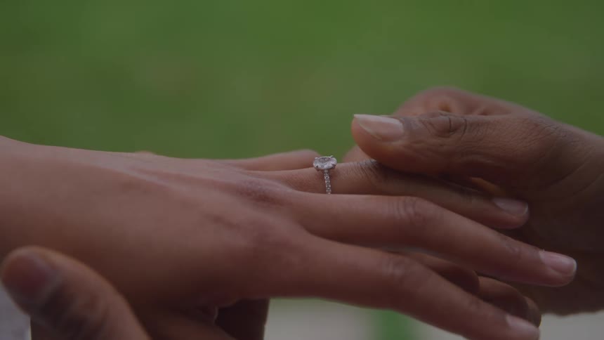 Hand places diamond ring on left ring finger in outdoor setting with blurred greenery