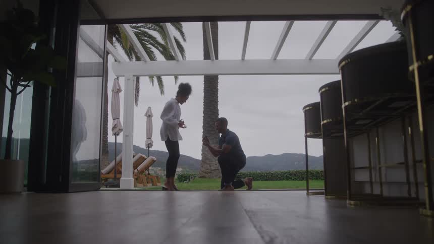A man proposes to a woman on a patio with a pergola and mountain view kneeling on one knee as she looks surprised framed by the interior of a house