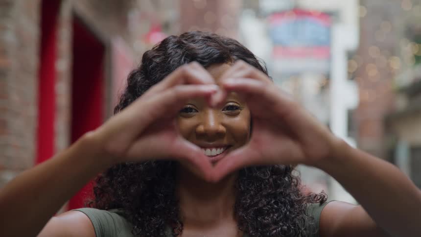 Curlyhaired person makes heart shape with hands in front of face in alleyway with lights