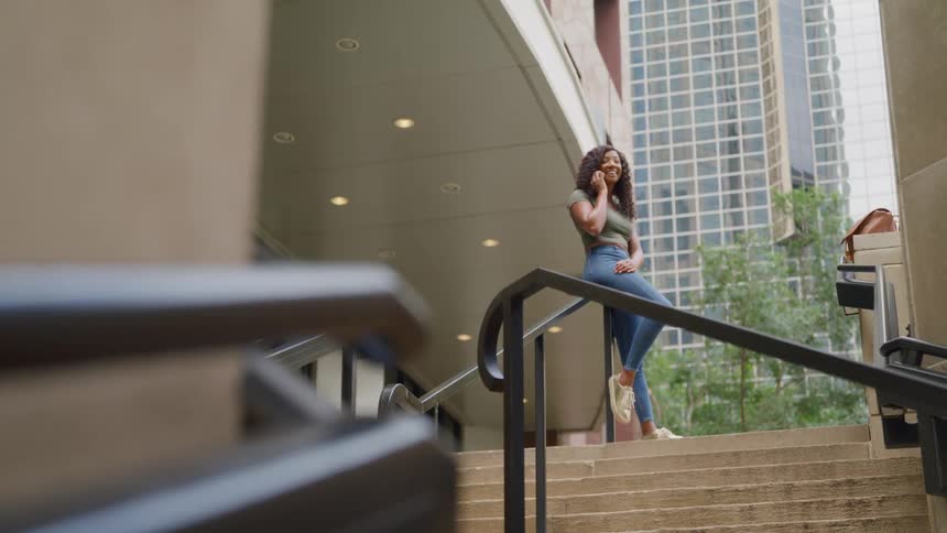 A young woman sits on the railing of a staircase in front of a building at midday happily making a phone call her excitement is visible as she smiles and chats soaking in the bright sunlight