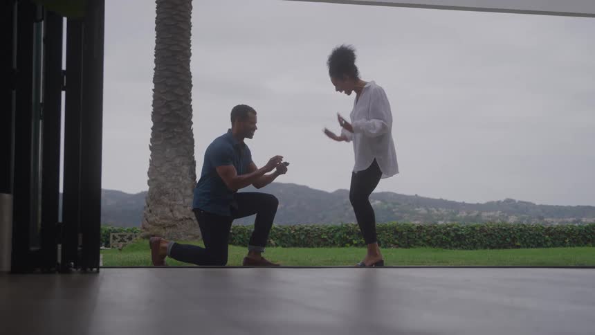 A man kneels down outdoors holding out a ring as he proposes marriage to a woman she stands before him surprised and emotional
