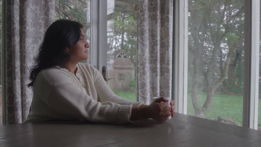 Woman sits at wooden table holding white mug with both hands in front of window with green view