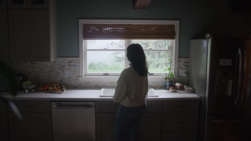 Woman stands in kitchen facing window surrounded by utensils and fruit in a calm modern setting