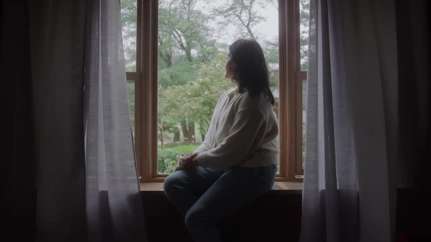 Woman sitting on the windowsill looks out the window toward the green outdoor area
