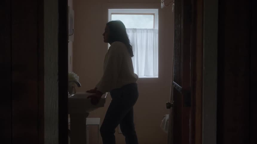 Person stands at sink in dim bathroom softly lit by light from window with white curtain